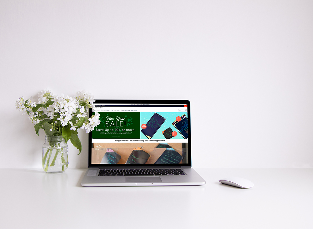Laptop displaying a New Year sale advertisement for writing solutions, alongside a vase of white flowers on a clean desk. The sale offers up to 20% off on various writing products, highlighting reusable writing and creativity solutions.