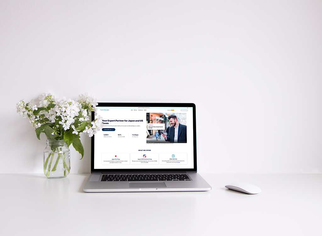 Laptop displaying a tax services website for Japan and US taxes, placed on a clean white desk with a vase of flowers, emphasizing a professional and organized workspace.