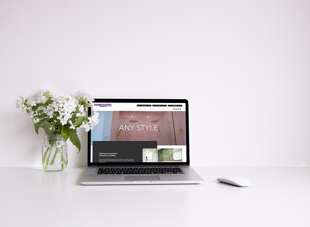 Laptop displaying a bathroom renovation website with the text "ANY STYLE" on the screen, accompanied by a vase of white flowers on a minimalistic desk.