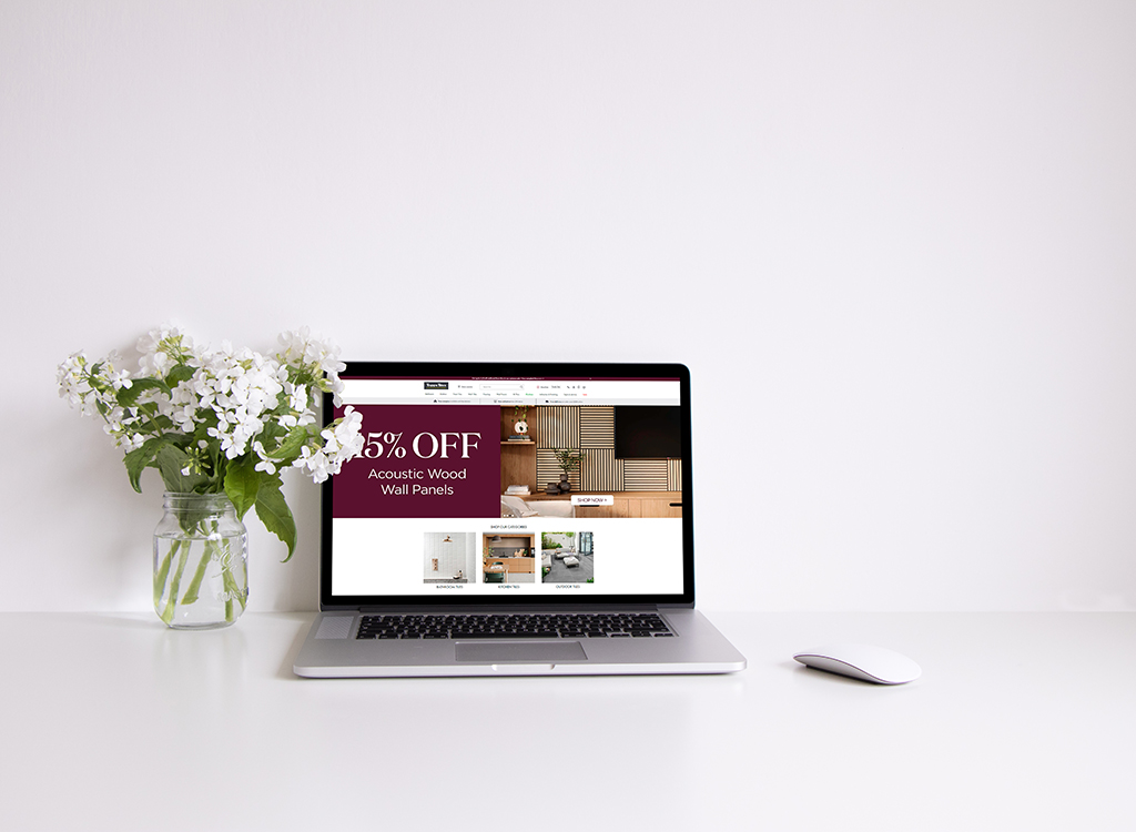 Laptop displaying a website offering 15% off on acoustic wood wall panels, accompanied by a vase of white flowers on a clean desk.