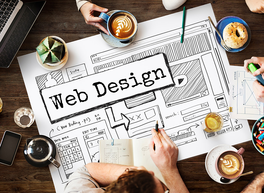 A top-down view of a creative workspace featuring a large sketch of a web design layout, coffee cups, and snacks, illustrating the process of website development and design brainstorming.